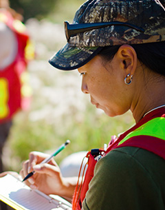 A person wearing a camouflage cap and a high-visibility safety vest writes notes on a clipboard outdoors during fieldwork.