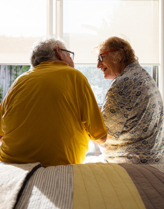 Two older adults sit on a bed facing each other, smiling and talking, with sunlight streaming through a window behind them.