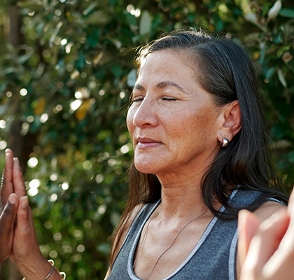 A woman with closed eyes holds her hands together outdoors, surrounded by greenery.