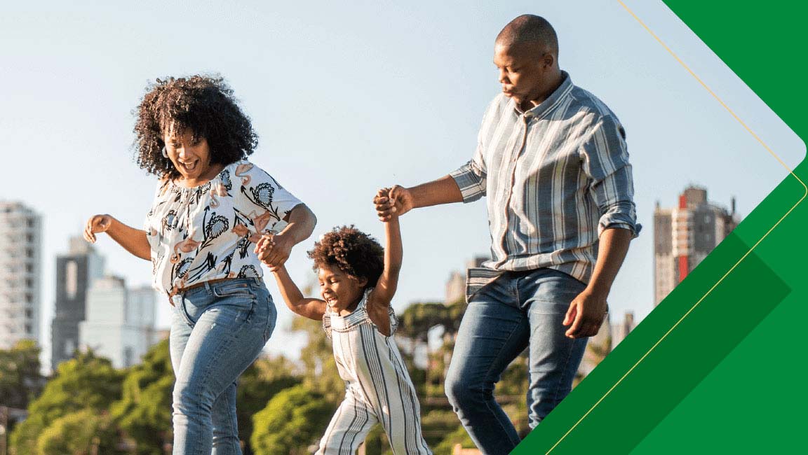 A family of three walks outdoors in a park, holding hands, with city buildings and trees in the background.