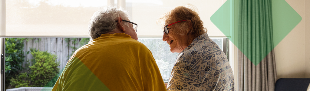 Two older adults sitting together indoors and talking by a window.